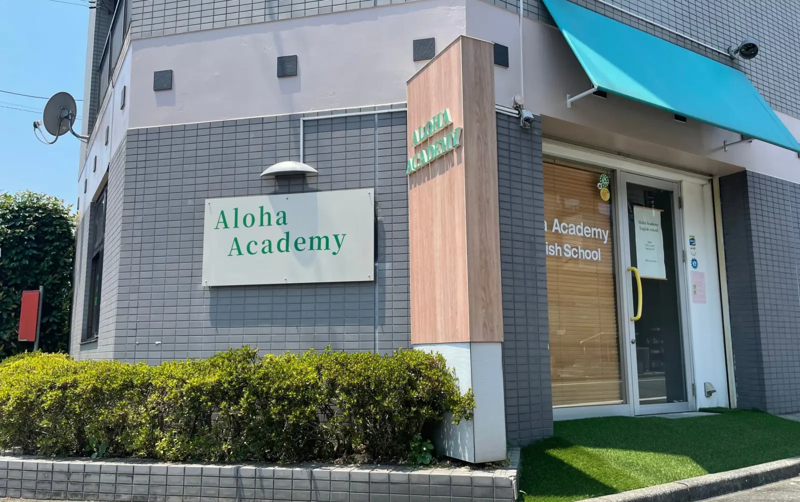グレータイル外壁の Aloha Academy 外観。ティール色の庇に大型看板、入口には人工芝。
